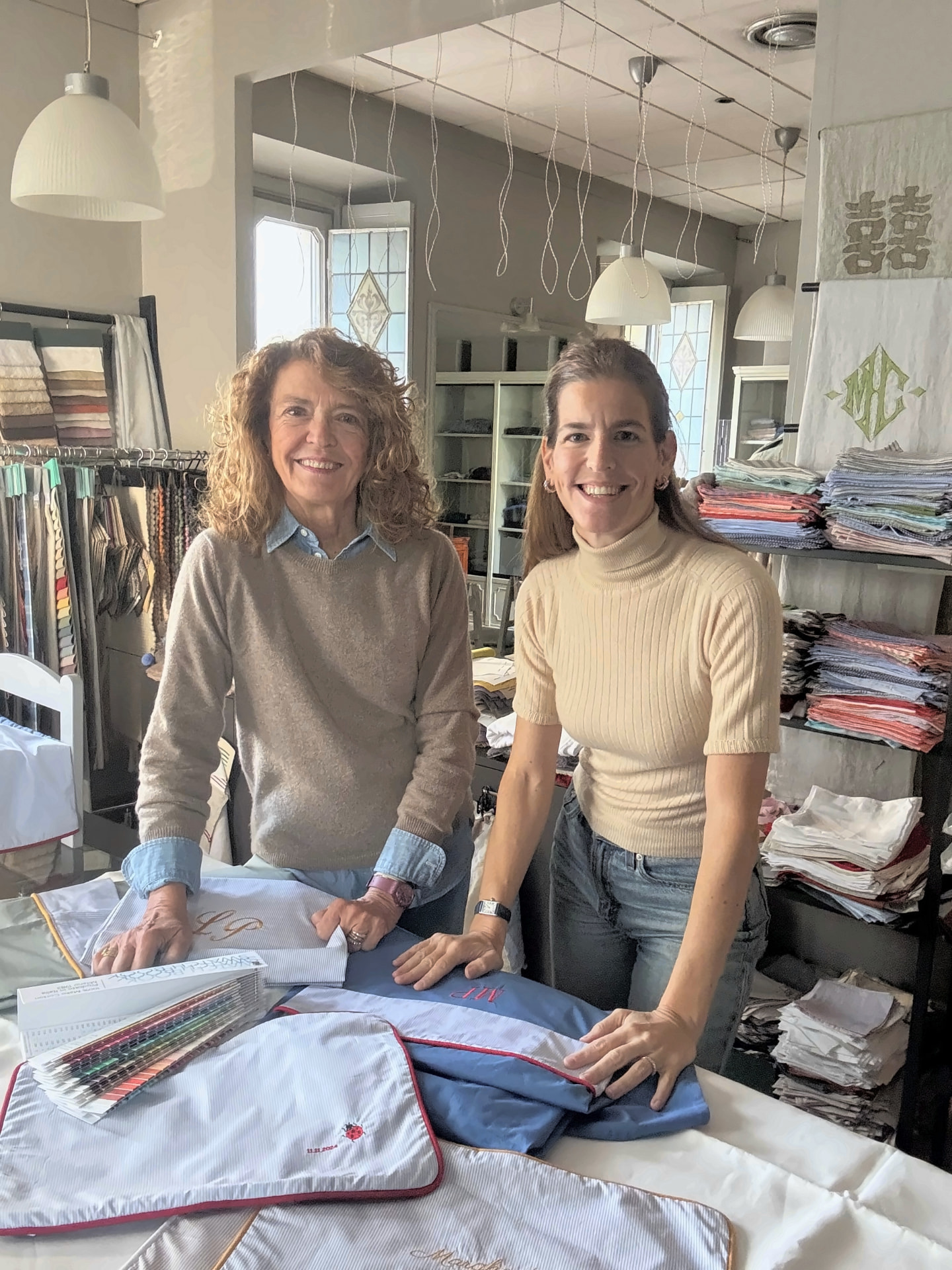 Two women standing behind a table of embroidered linens and fabric swatches in the MarinaC atelier in Milan