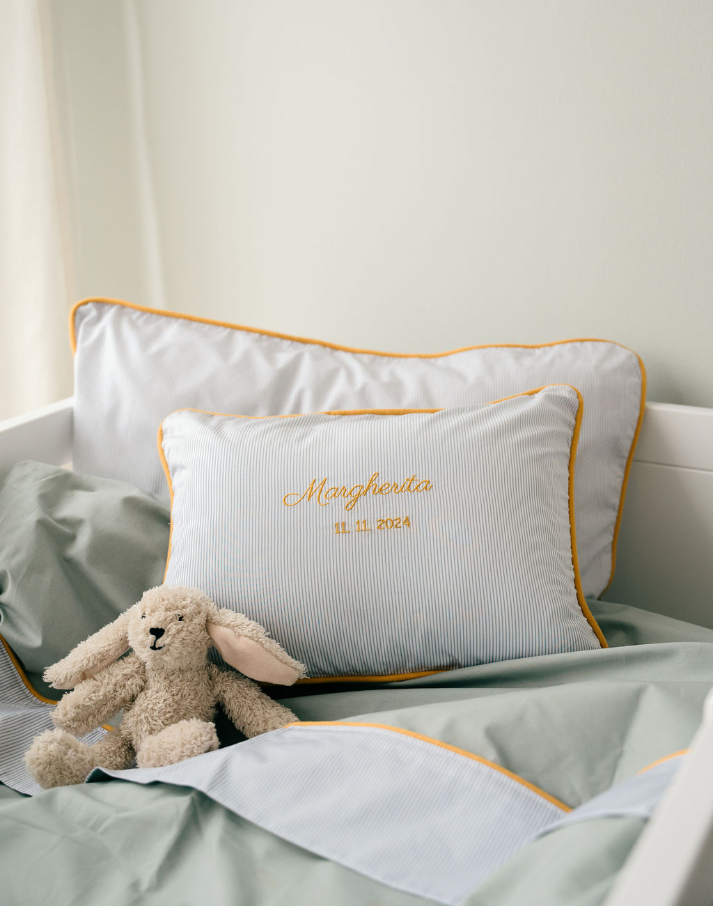 Leaf kids bedding on a white cot with pinstriped pillows, mustard piping, embroidered name and date, and a plush bunny