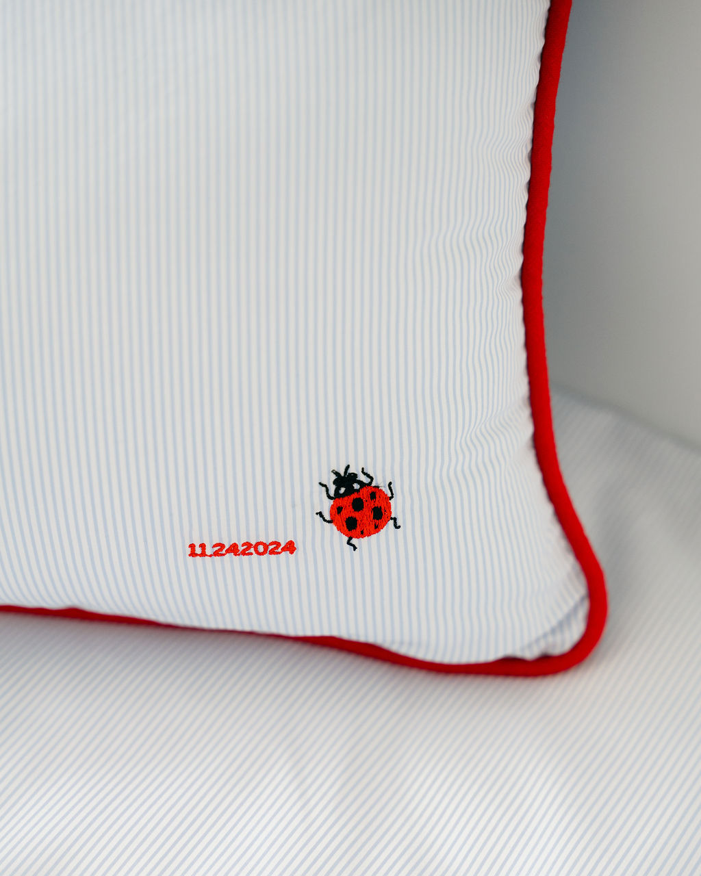 Blue pinstriped pillow with red piping, an embroidered ladybug motif and date, resting on matching striped bedding
